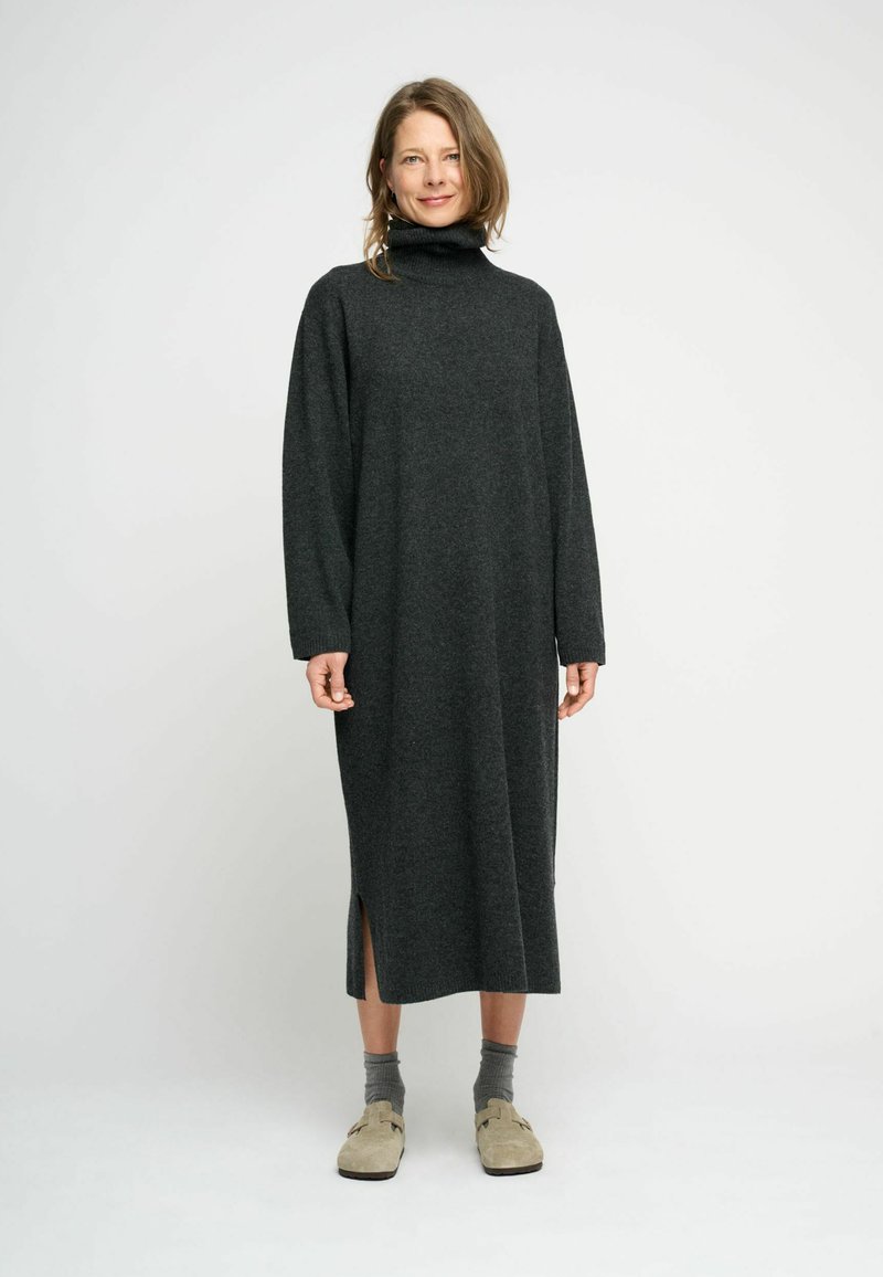Rochie din tricot cu mânecă lungă, de culoare gri închis, confecționată dintr-un material moale. Prezintă slițuri laterale și este asortată cu șosete gri și încălțăminte ușoară de culoare deschisă.
