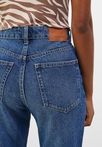 Jeans de mezclilla azul de tiro alto con costuras visibles, dos bolsillos traseros y una etiqueta de cuero marrón en la cintura. Tela texturizada.