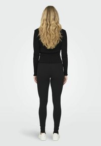 Must have musternööriga must pikkade varrukatega topp, millel on mustad legginsid, nähtav tagantpoolt, ja millel on sakilise äärega viimistlus ning valged tennisetossud.