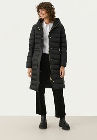 Zwarte pufferjas met een capuchon, voorzien van gouden hardware, een ritsluiting en horizontale gestikte secties. Gecombineerd met zwarte corduroy broek en laarzen.