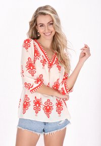 Blusa bianca con motivi floreali rossi e dettagli a righe, dotata di scollatura a V e maniche a tre quarti, abbinata a shorts in denim strappati.
