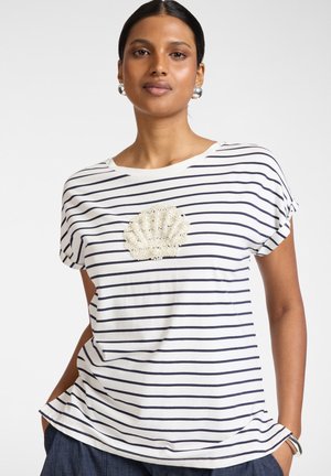 Mujer con camisa de rayas blancas y azul marino con diseño de concha adornado con perlas y pendientes de aro plateados, manos en los bolsillos.