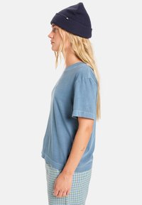 Kék pamut rövid ujjú póló, zöld kockás nadrággal és sötétkék kötött sapkával párosítva. Oldalnézeti kép.