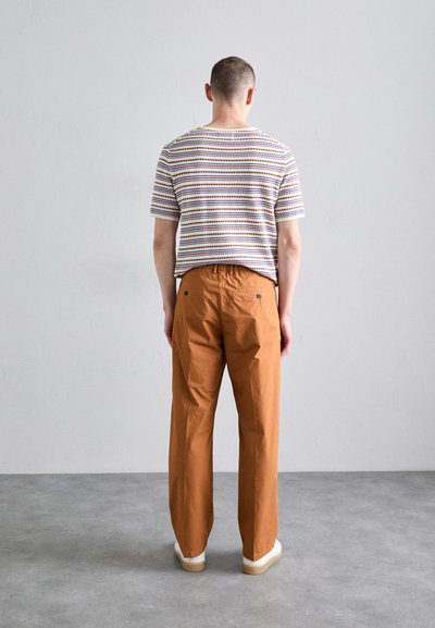 Chemise à manches courtes rayée en blanc, bleu et orange avec un pantalon orange à coupe décontractée, se tenant sur une surface grise.