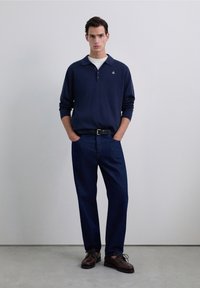 Polo din bumbac albastru închis cu mânecă lungă, cu un decolteu cu trei nasturi, asortat cu blugi de culoare albastru închis și pantofi cu șireturi maro. Design și texturi minimalist.