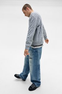 Lys grå strikket sweater med kabelmønster, slidte blå jeans og sorte sko. Casual outfit med lagdelte teksturer og synlig syning.