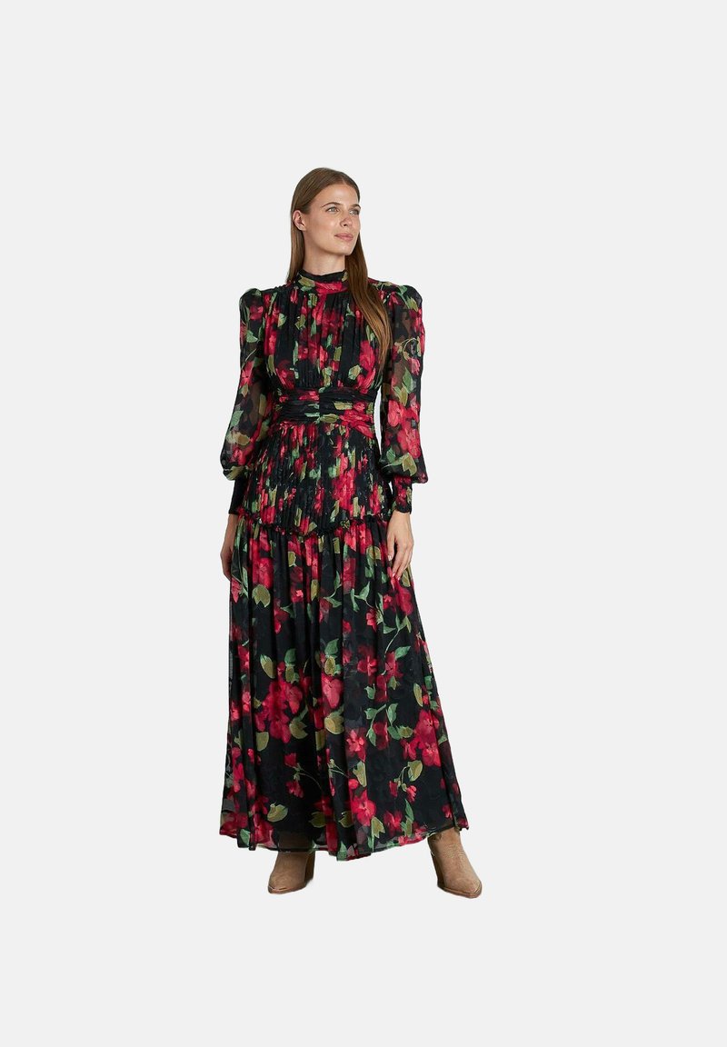 Vrouw in een lange zwarte jurk met rood en groen bloemenpatroon, hoge hals, poffende mouwen en tan enkelhoge laarzen, staande tegen een witte achtergrond.