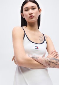 Νεαρή γυναίκα με μακριά σκούρα μαλλιά που φοράει λευκή φανέλα με μπλε περιθώριο και εμφανή λουλουδάτα και αφηρημένα τατουάζ στα χέρια και τον ώμο.