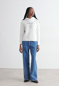 Hvid hætte sweatshirt med frontlogo, parret med blå bredbense denimjeans. Modellen står foran en neutral baggrund.