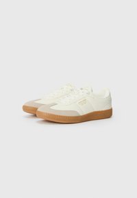 White sneakers with light grey accents, textured rubber sole, lace-up design, and branded detail in gold on the side.