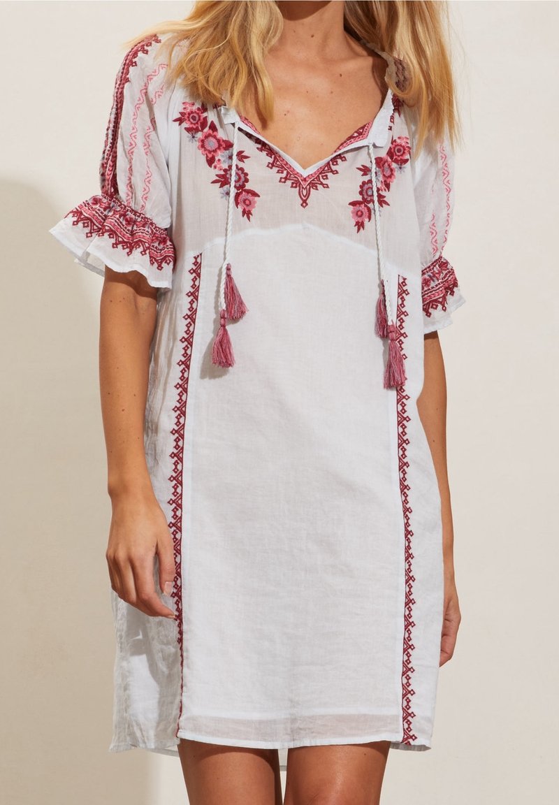 Robe blanche longueur genou avec broderie florale rouge, liens à pompons au col en V et manches courtes à volants portée par une personne.
