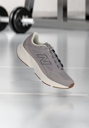 Scarpa da ginnastica grigia New Balance con suola bianca spessa sospesa sopra il pavimento della palestra, bilanciere sfocato e panca sullo sfondo.