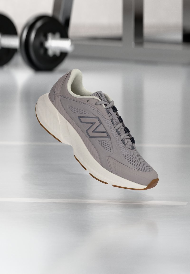 Γκρίζο αθλητικό παπούτσι New Balance με παχιά λευκή σόλα που αιωρείται πάνω από το πάτωμα του γυμναστηρίου, με θολό βάρος και πάγκο στο φόντο.