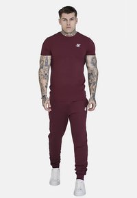 Bordowy komplet z bawełnianej koszulki i spodni dresowych, z dopasowanym krojem i logo na piersi, zestawiony z białymi trampkami.