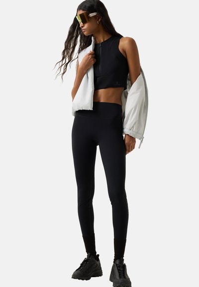 Kobieta ubrana w czarny krótki top i legginsy, z masywnymi czarnymi trampkami oraz białą kurtką zarzuconą na ramiona, pozuje na jednolitym tle.