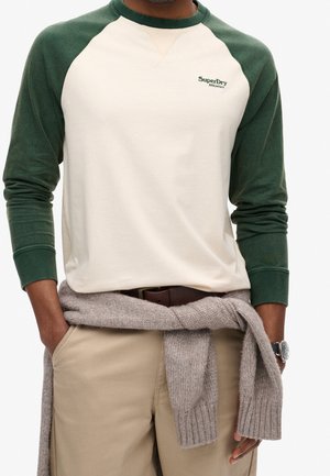 Elfenbeinfarbener Baseballshirt mit grünen Raglanärmeln und kleinem Logo. Beige Hose mit Gürtel, dazu ein hellgrauer Pullover um die Taille gebunden.