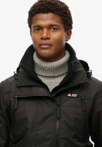 Sort zip-up jakke med tekstureret overflade, der har flere lynlås-lommer og en høj krave over en grå turtleneck sweater.