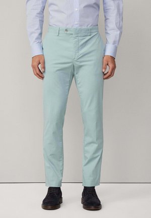 Pantalons en coton teal clair avec une taille plate, une fermeture éclair et un bouton unique. Porté avec une chemise à boutons bleu clair et des chaussures foncées.