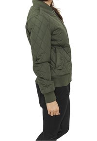 Una donna sta in posizione laterale, indossando una giacca bomber trapuntata verde e pantaloni neri. Ha i capelli lunghi e scuri e una posa rilassata.