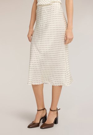 Frau trägt einen weißen Midirock mit schwarzen Punkten und braunen Leder-Blockabsatz-Sandaletten mit Knöchelriemen, steht vor hellem Hintergrund.