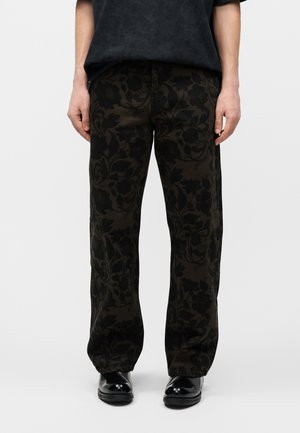 Άτομο που φορά μαύρο παντελόνι με σκούρα λουλουδάτα σχέδια και μαύρα γυαλιστερά παπούτσια, στέκεται μπροστά σε απλό ανοιχτόχρωμο φόντο.