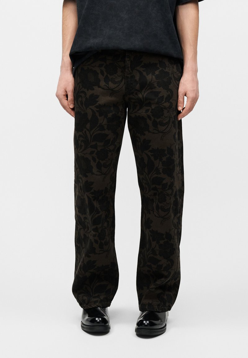 Άτομο που φορά μαύρο παντελόνι με σκούρα λουλουδάτα σχέδια και μαύρα γυαλιστερά παπούτσια, στέκεται μπροστά σε απλό ανοιχτόχρωμο φόντο.