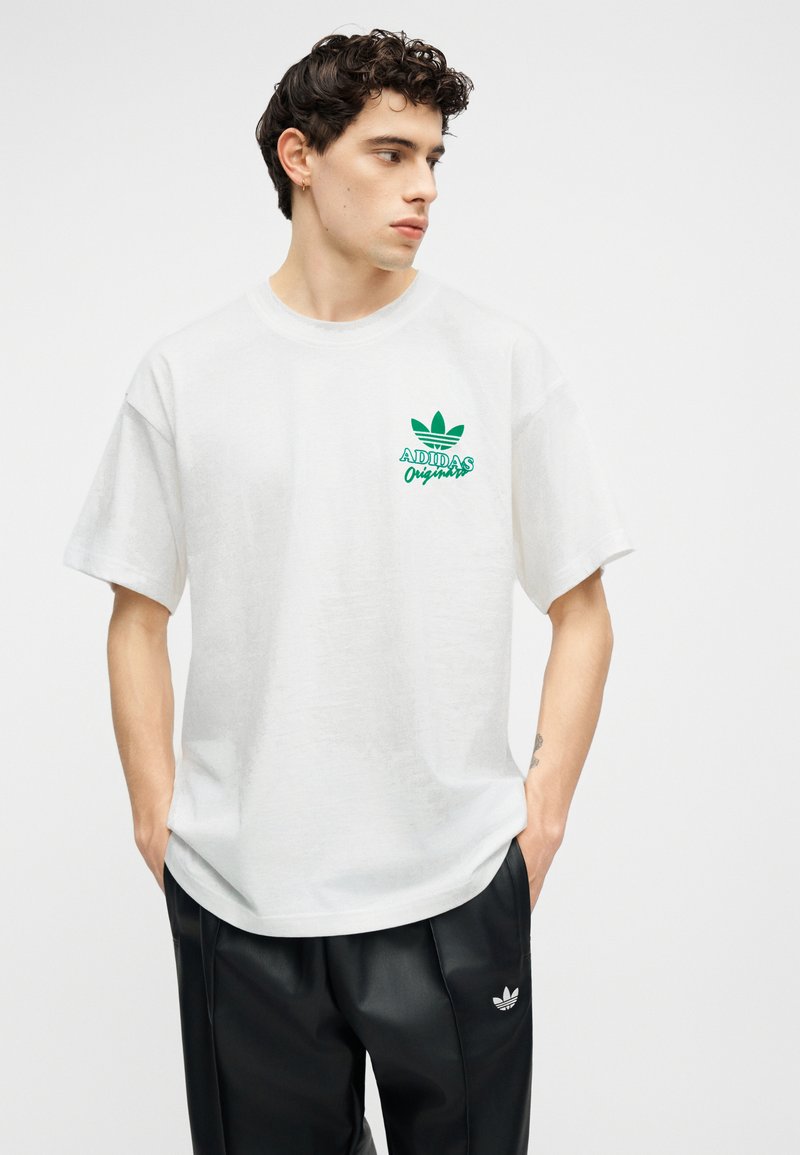 Jeune homme aux cheveux bouclés portant un t-shirt blanc Adidas Originals et un pantalon noir Adidas, regardant vers la gauche les mains dans les poches.