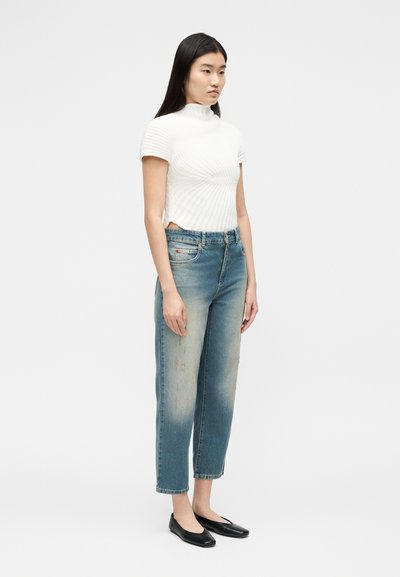 Femme debout portant un haut à col roulé côtelé blanc à manches courtes, un jean bleu délavé à jambes droites, et des chaussures plates noires.