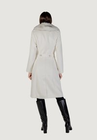 Manteau en laine blanche avec un col en fausse fourrure, de longueurmi-mi, au design ajusté et deux boutons à la taille, associé à des bottes noires jusqu'aux genoux.