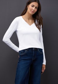 Weißes, langärmeliges V-Ausschnitt-Oberteil, kombiniert mit dunklen Jeans; glatte Textur mit figurbetontem Schnitt und subtilen Nähdetails.