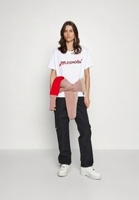 Hvid T-shirt med sort grafisk tekst, kombineret med sorte cargobukser. Rød og beige strikket sweater bundet om taljen. Hvide chunky sneakers.