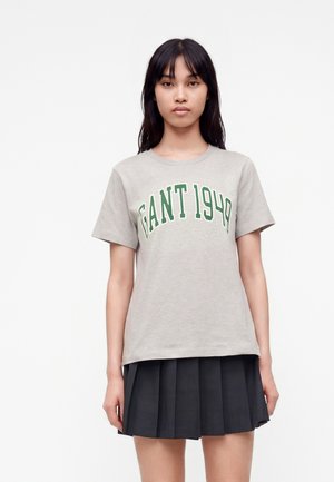 Jeune femme aux longs cheveux foncés portant un t-shirt gris clair avec un texte vert "GANT 1949" et une jupe plissée noire.