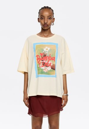 Camiseta oversized crema con un estampado gráfico colorido que presenta manos y flores, combinada con una falda sheer burdeos. Tejido de algodón suave.