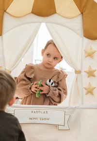 Žaidimų palapinė iš smėlio spalvos ir auksinio audinio, puošta trimatėmis žvaigždėmis ir vėliavnešiu su užrašu „FABELAB MINI SHOW“. Vaikas laiko minkštus žaislus.