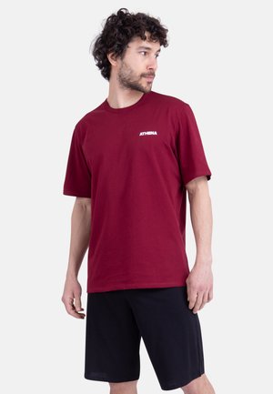 T-shirt en coton bordeaux à manches courtes et encolure ronde, avec le logo blanc "ATHENA" sur la poitrine gauche. Le modèle porte un short noir.