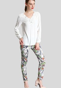 Blusa blanca de manga larga con detalles de encaje y escote en V, combinada con pantalones ajustados de estampado floral en colores vibrantes, llevados con zapatos de tacón beige.