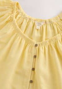 Blouse en lin jaune clair avec col froncé, petits boutons marron sur le devant et étiquette en tissu visible à l'intérieur du col.