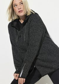 Felpa con zip nera e grigia a pois, con cappuccio e dettagli a costine in contrasto sulle maniche. Presenta una zip frontale e tasche.