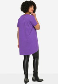 Paarse tuniek met een licht gebogen zoom, gecombineerd met zwarte, aansluitende leggings en gestepte zwarte enkellaarsjes. Glad textuur.