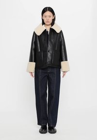 Zwarte leren jas met een crèmekleurige nepbont kraag en manchetten, met twee voorzakken en een ontspannen pasvorm, gedragen over donkerblauwe spijkerbroek.