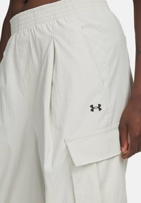Primer plano de una persona que lleva pantalones cargo blancos de Under Armour con cinturilla elástica y bolsillo lateral con solapa, con la mano descansando a un lado.