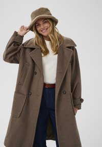 Brązowy wełniany płaszcz z dużym kołnierzem, dwoma przednimi kieszeniami i dwurzędowym zapięciem na guziki, w połączeniu z brązowym kapeluszem typu bucket.