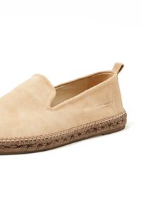 Chaussure slip-on en suède beige avec une semelle en jute tissée, un bout en forme d'amande et un petit onglet au talon. Marque en relief sur le côté.