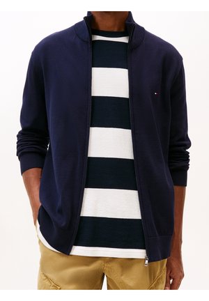 Hombre con chaqueta azul marino con cremallera sobre una camisa de rayas negras y blancas y pantalones beige, manos en los bolsillos, sobre un fondo claro.