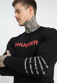 Sudadera negra de manga larga con "THUGGER" en rojo negrita en la parte delantera. Texto blanco con nombres de ciudades recorre ambas mangas.