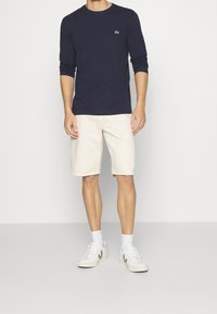 Man som bär en marinblå långärmad skjorta, beige knälånga shorts, vita strumpor och vita sneakers, stående mot en enfärgad bakgrund.