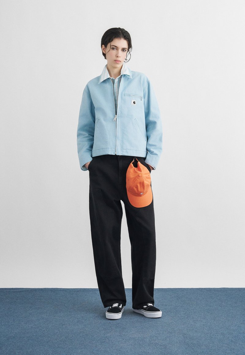 Jeune personne debout sur un tapis bleu portant une veste bleu clair, un pantalon large noir, des baskets noires, tenant une casquette orange à côté d'elle.