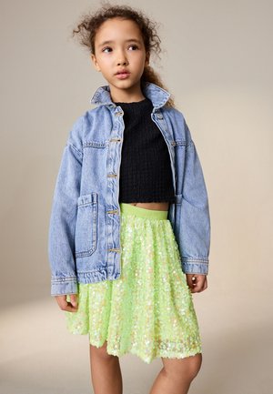 Noor tüdruk kannab sinist teksajopet musta topi ja erkrohelise litritega seelikuga, ta seisab ühtlase beeži taustaga.