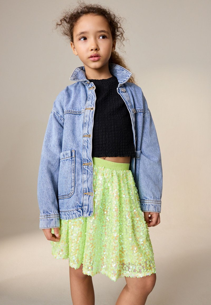 Jeune fille portant une veste en jean bleue sur un haut noir et une jupe à paillettes vert vif, debout devant un fond beige uni.