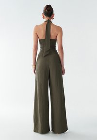 Une combinaison olive verte à jambes larges avec un col halter et un dos noué. Fabriquée en tissu texturé, elle présente un corsage ajusté et des pantalons fluides.